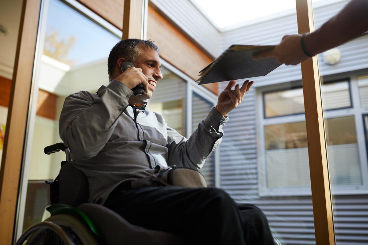 Auf dem ersten Foto sieht man einen Mann im Rollstuhl, der telefoniert und Unterlagen gereicht bekommt. Er trägt ein Hemd und sieht sehr beschäftigt aus.