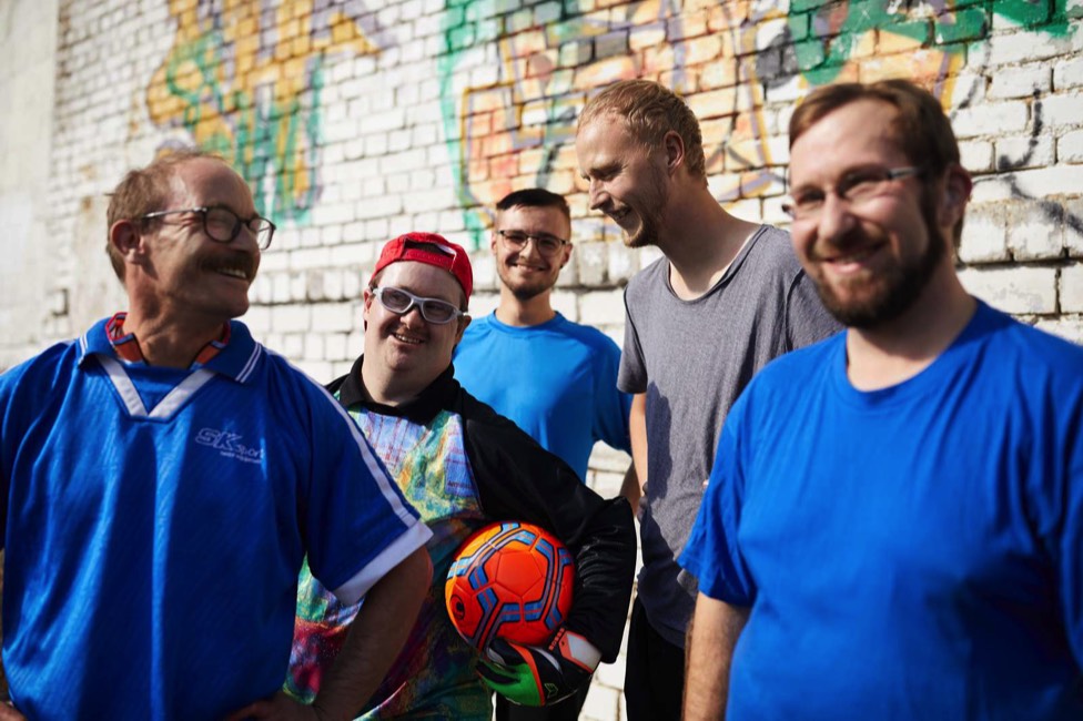 Auf dem 8. Bild sieht man Freiwillige und Menschen mit Beeinträchtigung draußen beim Sport, ein Mann hält einen Ball unter dem Arm.