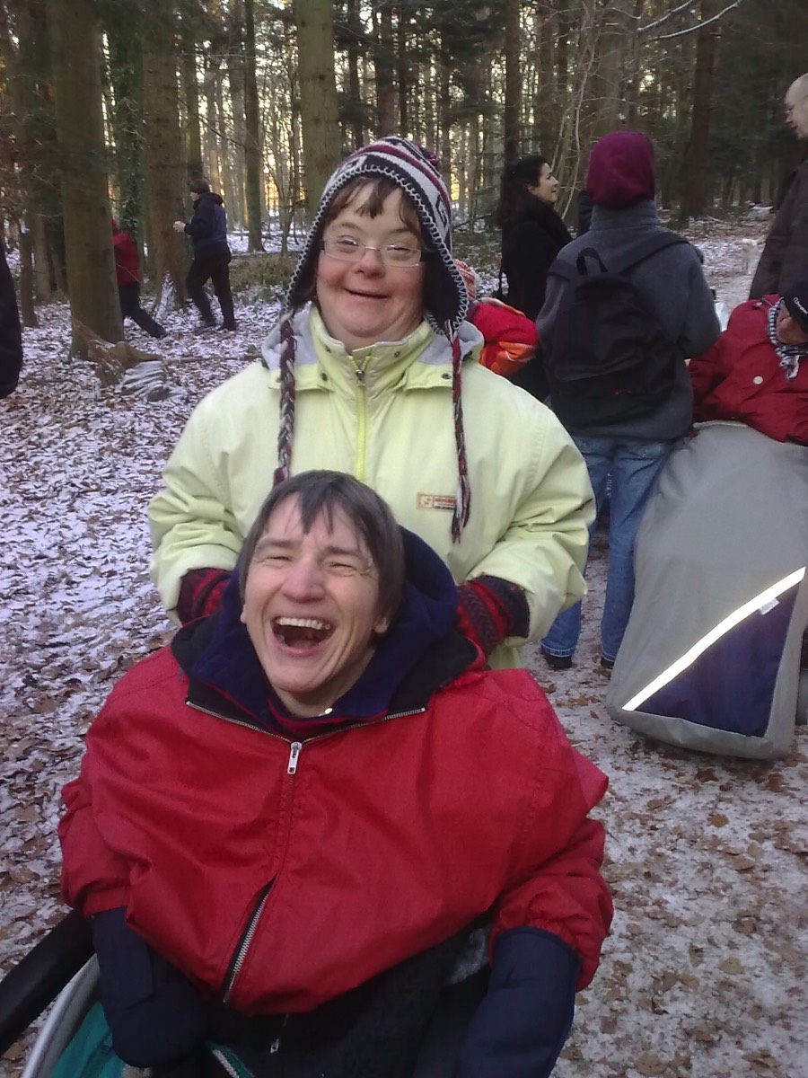 Auf dem 2. Bild sieht man eine Frau mit Trisomie 21, die eine andere Frau im Rollstuhl durch den Wald schiebt. Beide sind warm angezogen und lachen, es liegt etwas Schnee.