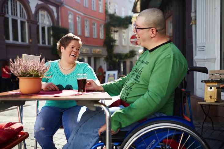 Auf dem 3. Bild sieht man eine Freiwillige und einen Mann im Rollstuhl drauÃŸen in einem CafÃ©.