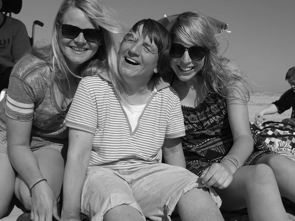 Auf dem Bild sieht man zwei Frauen ohne BeeintrÃ¤chtigung und eine Frau mit BeeintrÃ¤chtigung. Sie sitzen am Strand und lachen zusammen.