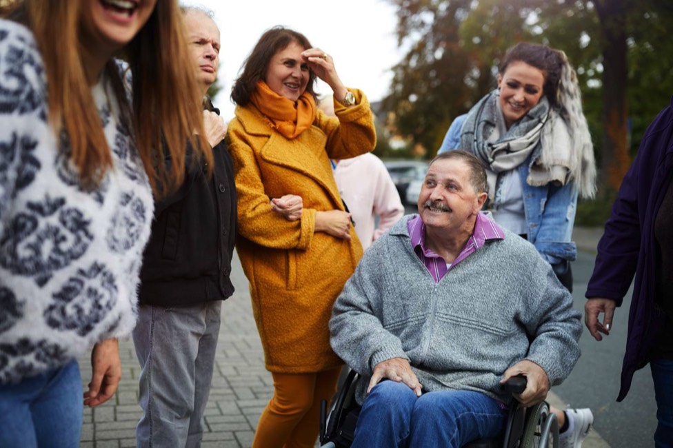 Auf dem 7. Bild sieht man eine Gruppe Freiwilliger und einen Mann im Rollstuhl, die zusammen unterwegs sind und Spaß haben.