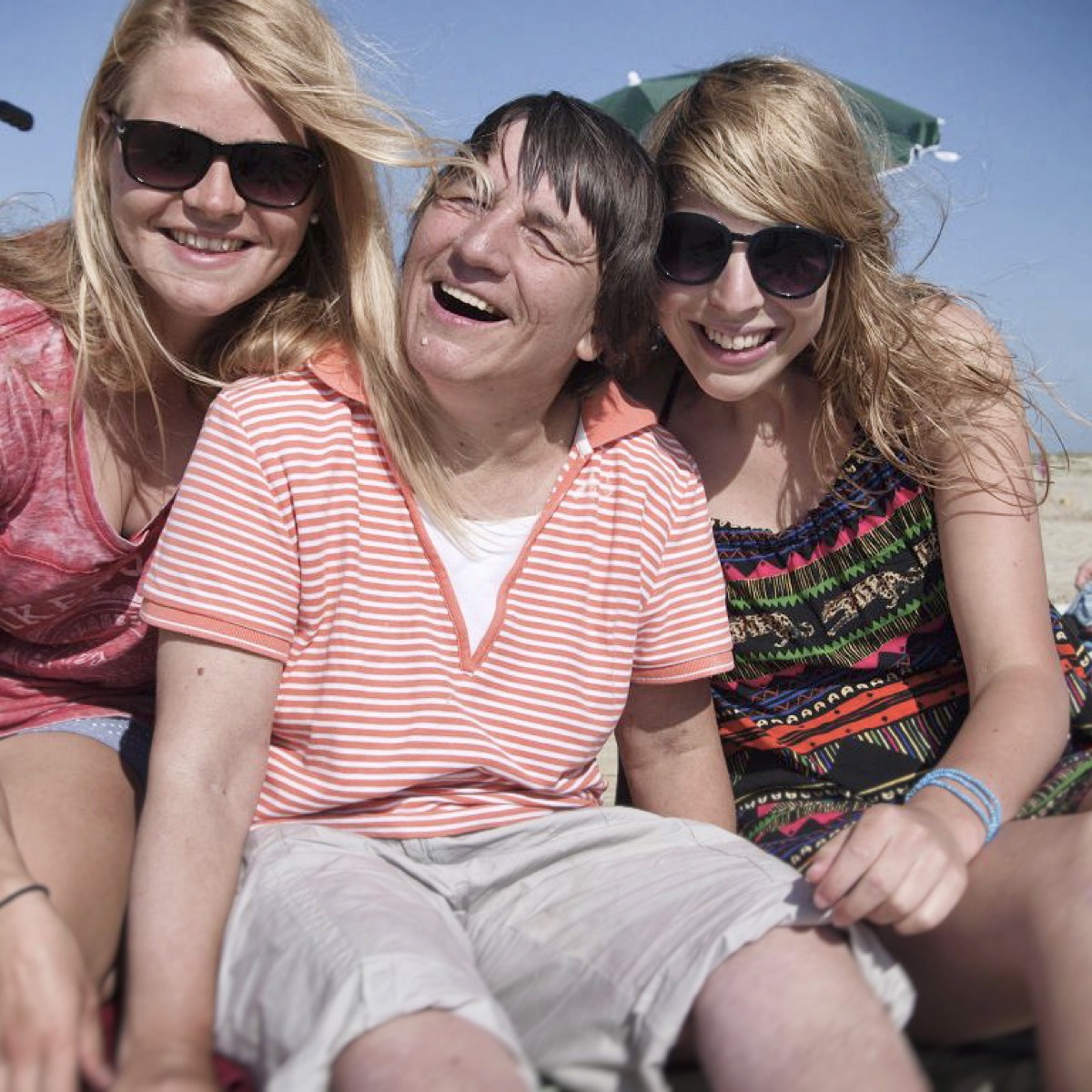 Auf dem 1. Bild sieht man zwei Frauen ohne Beeinträchtigung und eine Frau mit Beeinträchtigung. Sie sitzen am Strand und lachen zusammen.