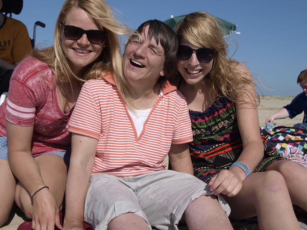 Auf dem 1. Bild sieht man zwei Frauen ohne BeeintrÃ¤chtigung und eine Frau mit BeeintrÃ¤chtigung. Sie sitzen am Strand und lachen zusammen.